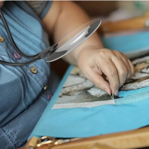 Close-up of hand stitching fabric under magnifying glass.